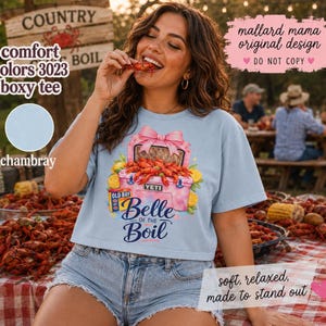 Puede incluir: Camiseta boxy azul claro con un gráfico de una nevera rosa llena de cangrejos de río, limones y un lazo rosa. La camiseta tiene el texto "Belle of the Boil" y "Yeti". La camiseta también tiene el texto "comfort colors 3023 boxy tee" y "chambray".