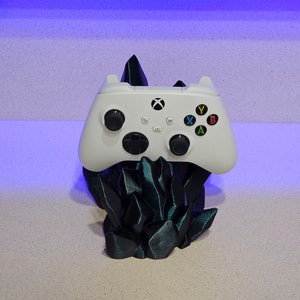 May include: A white Xbox controller sits on a black, crystal-shaped stand. The controller has black buttons and a green Xbox logo.