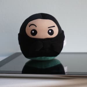 May include: A black and green plush toy shaped like a ninja with a black mask and white eyes. The toy is sitting on a black tablet.