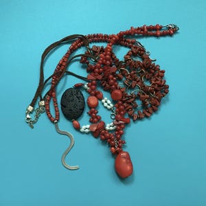 Puede incluir: Una colección de joyas que incluye varios collares con cuentas de coral rojo, pequeñas perlas blancas y una cuenta de piedra de lava negra porosa. Un collar incluye un cordón de cuero marrón y un componente colgante de metal plateado ondulado.