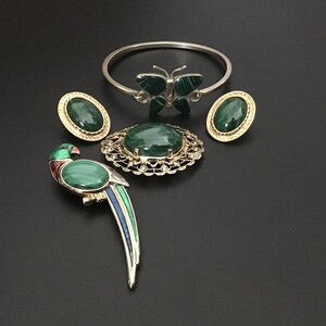 Puede incluir: Una colección de joyas que incluye un brazalete de mariposa plateado con alas de malaquita verde, dos pendientes ovalados de malaquita, un broche redondo de malaquita con borde de filigrana y un broche con forma de loro en esmalte verde, azul y rojo.