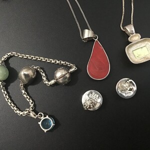 Puede incluir: Una colecci&oacute;n de collares y abalorios de plata sobre una superficie negra. Los collares presentan colgantes en rojo y una forma cuadrada con un centro verde. Tambi&eacute;n hay una pulsera con cuentas verdes y plateadas, junto con dos peque&ntilde;os abalorios redondos.