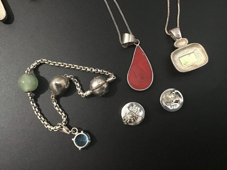 Puede incluir: Una colecci&oacute;n de collares y abalorios de plata sobre una superficie negra. Los collares presentan colgantes en rojo y una forma cuadrada con un centro verde. Tambi&eacute;n hay una pulsera con cuentas verdes y plateadas, junto con dos peque&ntilde;os abalorios redondos.