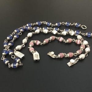 Puede incluir: Una colecci&oacute;n de collares y pulseras de cuentas en tonos azules, blancos y rosas. La joyer&iacute;a presenta peque&ntilde;as cuentas redondas ensartadas con eslabones y cierres de metal plateado. Las piezas est&aacute;n dispuestas sobre una superficie oscura.