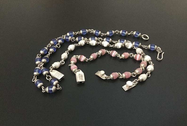 Puede incluir: Una colecci&oacute;n de collares y pulseras de cuentas en tonos azules, blancos y rosas. La joyer&iacute;a presenta peque&ntilde;as cuentas redondas ensartadas con eslabones y cierres de metal plateado. Las piezas est&aacute;n dispuestas sobre una superficie oscura.