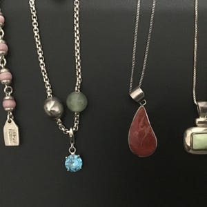 Puede incluir: Una colecci&oacute;n de collares con varios dise&ntilde;os de colgantes. Los collares incluyen hilos de cuentas con cuentas rosas, una cadena con una gema azul y colgantes con piedras rojas y verdes. Las joyas se exhiben sobre un fondo negro.