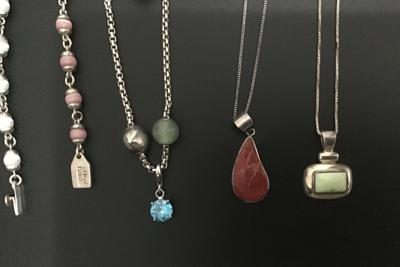 Puede incluir: Una colecci&oacute;n de collares con varios dise&ntilde;os de colgantes. Los collares incluyen hilos de cuentas con cuentas rosas, una cadena con una gema azul y colgantes con piedras rojas y verdes. Las joyas se exhiben sobre un fondo negro.