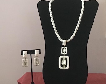 Puede incluir: Un collar de cuerda blanco con un colgante blanco que presenta tres formas cuadradas apiladas con diamantes de imitación transparentes. También se incluyen pendientes blancos a juego con diamantes de imitación transparentes.