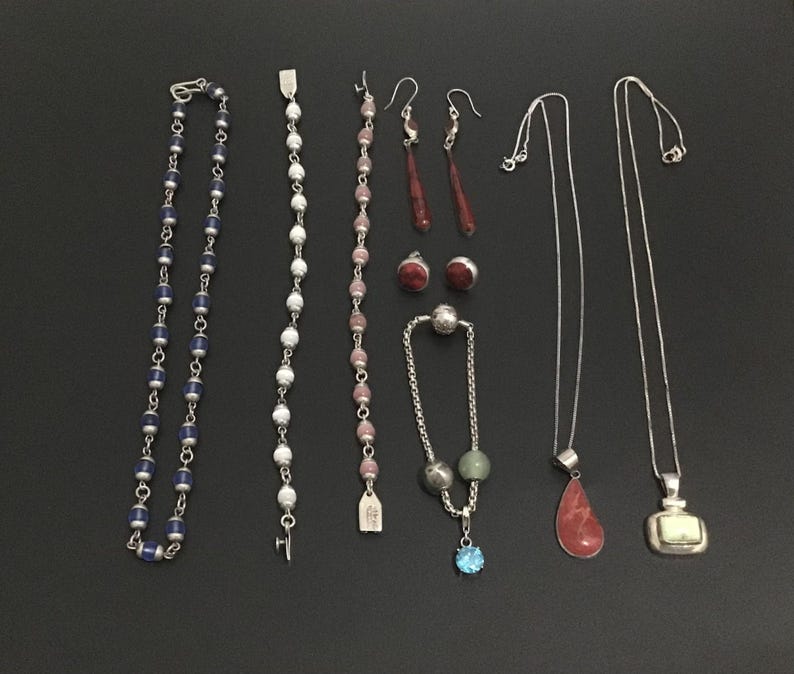 Puede incluir: Una variedad de piezas de joyer&iacute;a, que incluyen collares, pulseras y aretes, exhibidas sobre una superficie negra. La joyer&iacute;a presenta varias piedras preciosas, cuentas y cadenas de plata. La colecci&oacute;n incluye un collar de cuentas azules, una pulsera de cuentas blancas y aretes rojos.
