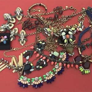 Puede incluir: Una colección de varios collares y pendientes con piedras preciosas de colores y metal dorado. La joyería incluye collares llamativos, cadenas delicadas y pendientes de botón. Las piezas están dispuestas sobre una superficie roja.
