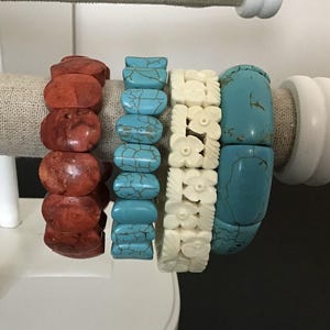 Puede incluir: Cuatro pulseras elásticas en un soporte. La primera es de cuentas ovaladas de color marrón rojizo, la segunda de cuentas azul turquesa con vetas oscuras, la tercera de cuentas color crema talladas con flores y la cuarta de cuentas azules lisas.