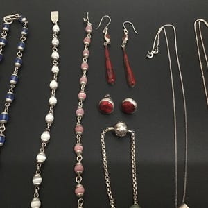 Puede incluir: Un surtido de piezas de joyer&iacute;a que incluye collares, aretes y una pulsera. Los collares presentan cuentas azules y blancas, y delicadas cadenas de plata. Los aretes incluyen dise&ntilde;os rojos de l&aacute;grima y de bot&oacute;n redondo. La joyer&iacute;a se exhibe sobre una superficie negra.