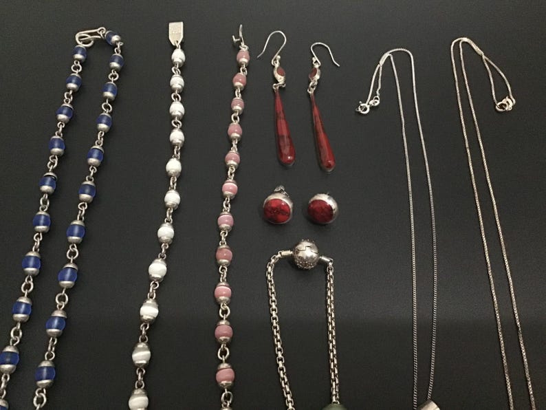 Puede incluir: Un surtido de piezas de joyer&iacute;a que incluye collares, aretes y una pulsera. Los collares presentan cuentas azules y blancas, y delicadas cadenas de plata. Los aretes incluyen dise&ntilde;os rojos de l&aacute;grima y de bot&oacute;n redondo. La joyer&iacute;a se exhibe sobre una superficie negra.