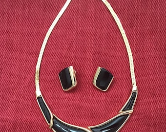 May include: A gold-toned necklace and matching earrings set. The necklace features a black enamel design with gold accents. The earrings are square-shaped with black enamel and gold trim. The jewelry set is displayed on a red background.