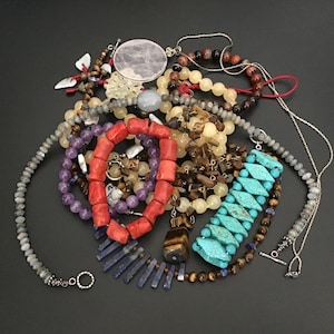 Puede incluir: Una colección de joyas variadas que incluye una pulsera de cuentas de coral rojo, una pulsera de cuentas turquesas en forma de diamante, un collar de cuentas de amatista morada, un collar de cuentas grises facetadas y otros collares.
