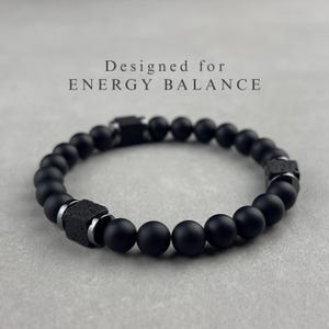 Könnte beinhalten: Ein Armband aus mattschwarzen, kugelförmigen Perlen, unterbrochen von zwei schwarzen, würfelförmigen, texturierten Perlen und silberfarbenen Metallzwischenstücken. Der Text "Designed for ENERGY BALANCE" steht über dem Armband.