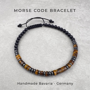 Op de afbeelding: Een Morsecode-armband met een herhalend patroon van matzwarte ronde kralen, bruine cilindrische tijgeroogkralen en kleine metallic grijze tussenringen. De armband is voorzien van een verstelbare zwarte koordsluiting.