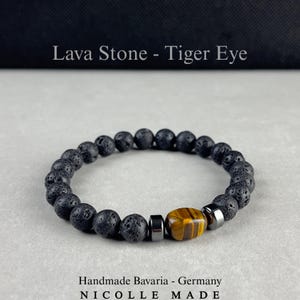 Könnte beinhalten: Ein Armband aus runden, schwarzen, porösen Lavasteinperlen, zwei metallischen Zwischenelementen und einer rechteckigen Tigerauge-Perle mit braunen und goldenen Streifen. Der Text Lava Stone - Tiger Eye, Handmade Bavaria - Germany, NICOLLE MADE ist sichtbar.