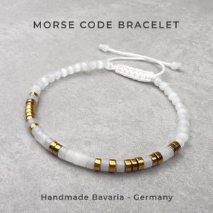 Könnte beinhalten: Ein Morsecode-Armband mit einer Reihe weißer Perlen, die mit kleinen, zylindrischen goldfarbenen Metallperlen durchsetzt sind. Das Armband verfügt über einen verstellbaren weißen Kordelverschluss mit zwei weißen Perlen an den Enden.