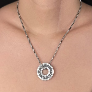 Peut inclure: Un pendentif circulaire de couleur argentée orné de caractères runiques gravés sur son bord extérieur, suspendu à une chaîne à maillons carrés assortie. Le pendentif présente une finition métallique lisse avec des lettres sombres.