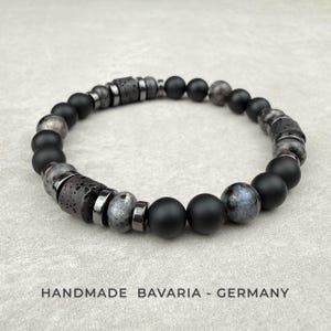 Peut inclure: Un bracelet de perles composé de perles rondes noir mat, de perles en pierre de lave grise poreuse et texturée, et de perles grises marbrées lisses, avec des entretoises métalliques argentées. Le texte HANDMADE BAVARIA - GERMANY est inscrit.