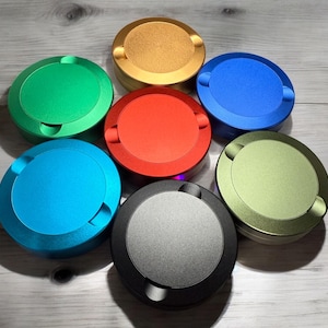 May include: A collection of seven round, metallic ashtrays in various colors: green, gold, blue, red, light green, light blue, and black. Each ashtray has a circular design with a recessed area for resting cigarettes. The ashtrays are arranged on a light wooden surface.