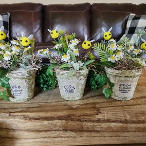May include: Three decorative flower pots with white daisies, green foliage, and yellow bee accents. The pots have a distressed finish and feature inspirational text. The arrangement sits on a wooden tray.