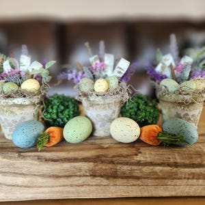 May include: Easter-themed decorative arrangement featuring three small pots with speckled eggs, faux greenery, and lavender. The arrangement includes speckled eggs in blue, green, and white, along with orange carrot decorations, all displayed on a wooden tray.