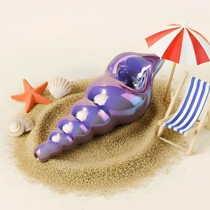 Puede incluir: Una figura de concha iridiscente púrpura descansa sobre arena, acompañada de una sombrilla de playa en miniatura y una silla de playa a rayas. Conchas marinas adicionales y una estrella de mar están dispersas, creando un tema de playa.