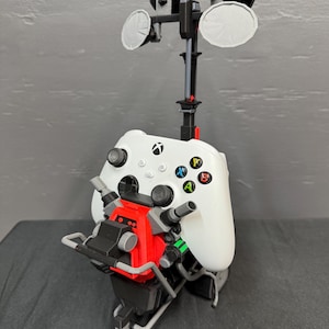 May include: A white Xbox wireless controller mounted on a black and red 3D-printed stand. The stand features a tall vertical pole with three circular satellite-like dishes at the top and a mechanical base with gray accents and red detailing.