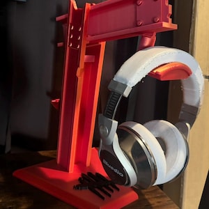 May include: A red 3D printed headphone stand with a metal beam design. The stand has a base and a vertical support with a horizontal arm that holds the headphones. The headphones are black with white earcups and the brand name "Bluedio" is visible on the left earcup.