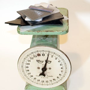 May include: An antique green Way Rite scale with a white face and black numbers. The scale reads zero pounds. A pile of black and gray rectangular objects sits on the scale's platform.
