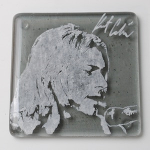 May include: A gray glass coaster with a white silhouette of a person's face and the signature "Kurt Cobain" in white.