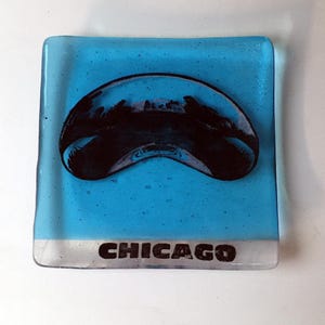 May include: A blue glass coaster with a black silhouette of the Chicago skyline in the shape of a jelly bean. The word "CHICAGO" is printed in black on the bottom edge of the coaster.