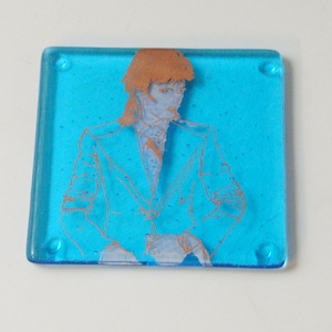 May include: A blue glass coaster with a white outline of a person wearing a suit. The person's hair is a copper color.