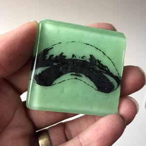 May include: A green glass tile with a black and white abstract design. The design resembles a wave or a mountain range.