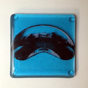 May include: A blue glass coaster with a black and white abstract design. The design resembles a curved shape with a textured surface.
