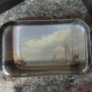 May include: Rectangular glass tray with a silver trim. The tray features a seascape painting with sailboats on the water under a cloudy sky. The painting has muted tones of blue, beige, and brown.