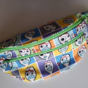 May include: A green and multi-colored fabric fanny pack with a repeating pattern of cartoon faces. The faces are white with black eyes and red mouths. The fanny pack has two green zippers.