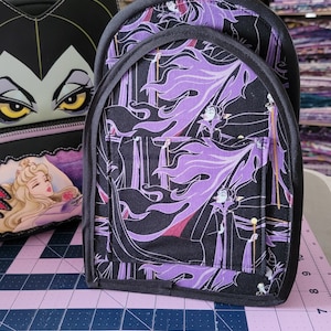 May include: A black and purple fabric backpack with a Maleficent print. The backpack has a zipper closure and a small pocket on the front.