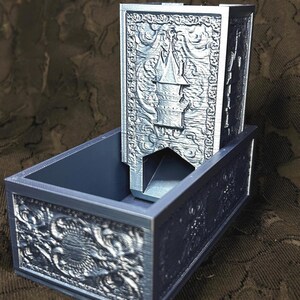 Puede incluir: Un juego de torre y bandeja para dados de color azul oscuro. La torre presenta un diseño detallado de castillo, mientras que la bandeja tiene intrincados diseños florales y de dragones. El juego está hecho de un material sólido y está diseñado para juegos de mesa.