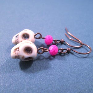 May include: A pair of dangle earrings featuring off-white skull-shaped beads and bright pink faceted round beads. The components are connected by copper-toned metal jump rings and suspended from matching copper-toned metal fishhook ear wires.