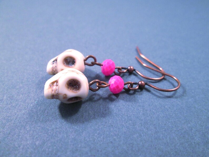 May include: A pair of dangle earrings featuring off-white skull-shaped beads and bright pink faceted round beads. The components are connected by copper-toned metal jump rings and suspended from matching copper-toned metal fishhook ear wires.