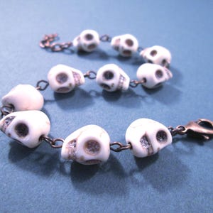 May include: A bracelet featuring a series of white, skull-shaped beads with dark, recessed eye sockets and nasal cavities. The beads are connected by copper-toned metal jump rings and include a copper-toned lobster claw clasp at one end.