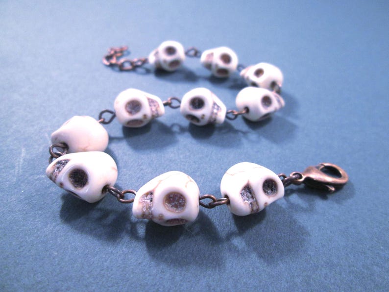 May include: A bracelet featuring a series of white, skull-shaped beads with dark, recessed eye sockets and nasal cavities. The beads are connected by copper-toned metal jump rings and include a copper-toned lobster claw clasp at one end.