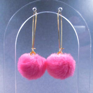 May include: A pair of dangling earrings featuring bright pink, fluffy pom-poms. The earrings have long, thin gold-colored wires that loop through the ear. The pom-poms are round and soft, creating a playful and eye-catching accessory.