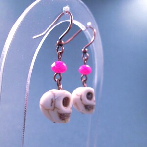 May include: A pair of drop earrings featuring white skull-shaped beads with brown veining, accented by small, faceted hot pink beads. The skulls are suspended from dark metal ear wires by short metal links, creating a dangling design.