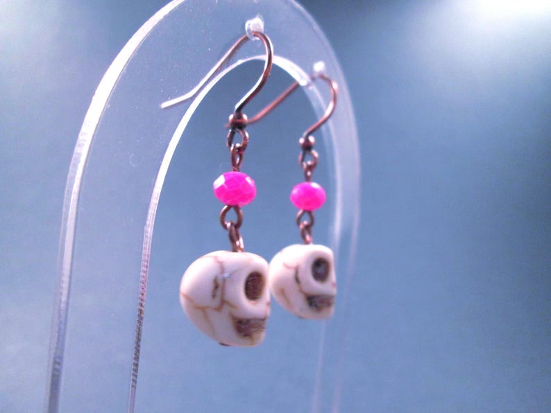 May include: A pair of drop earrings featuring white skull-shaped beads with brown veining, accented by small, faceted hot pink beads. The skulls are suspended from dark metal ear wires by short metal links, creating a dangling design.