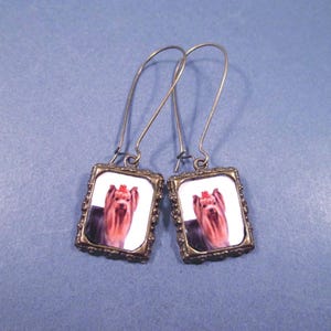 May include: A pair of dangle earrings featuring rectangular pendants with a portrait of a Yorkshire Terrier. The dog has long, flowing hair and a red bow. The pendants are framed in ornate, antique-style metal.