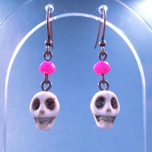 May include: A pair of drop earrings featuring white skull-shaped beads with dark eye sockets and teeth, accented by bright pink faceted beads. The skulls are suspended from copper-toned metal ear wires, creating a distinct contrast between the light skull beads and the vibrant pink accents.